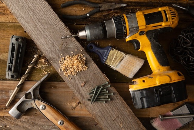 A variety of tools, including a hammer and cordless drill, sit on top of a table made of wood boards